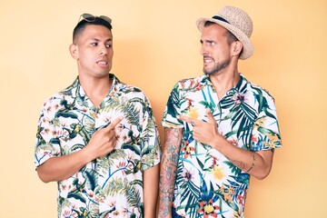 Young gay couple of two men wearing summer hat and hawaiian shirt pointing aside worried and nervous with forefinger, concerned and surprised expression