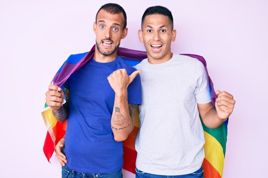 Young gay couple of two men holding rainbow lgbtq flag together pointing thumb up to the side smiling happy with open mouth
