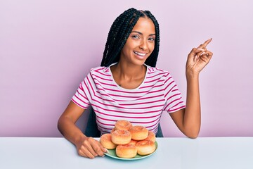 Beautiful hispanic woman eating doughnut for breakfast smiling happy pointing with hand and finger to the side