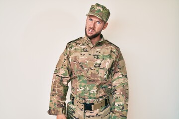 Young caucasian man wearing camouflage army uniform in shock face, looking skeptical and sarcastic, surprised with open mouth