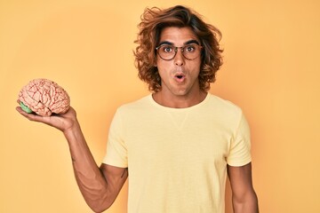 Young hispanic man holding brain scared and amazed with open mouth for surprise, disbelief face