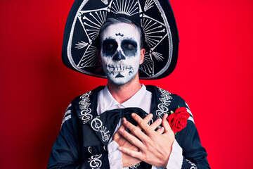 Young man wearing mexican day of the dead costume over red smiling with hands on chest with closed eyes and grateful gesture on face. health concept.