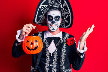 Young man wearing mexican day of the dead costume holding pumpkin celebrating victory with happy smile and winner expression with raised hands