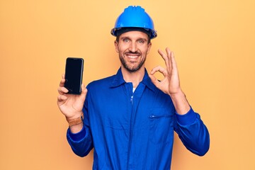 Young handsome worker man wearing uniform and hardhat holding smartphone showing screen doing ok sign with fingers, smiling friendly gesturing excellent symbol