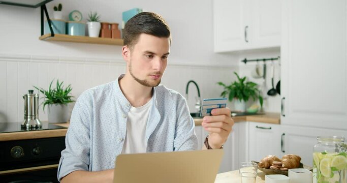 Young Man Pays Online by Credit Card using Laptop in Kitchen. Customer Buys or makes Purchase at Home in Web Store and Enjoys Shopping via Internet. E-banking and E-commerce Concept. 4K Medium Shot