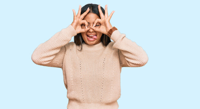 Young latin girl wearing wool winter sweater doing ok gesture like binoculars sticking tongue out, eyes looking through fingers. crazy expression.