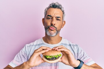 Middle age grey-haired man eating a tasty classic burger looking at the camera blowing a kiss being lovely and sexy. love expression.