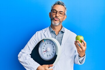Middle age grey-haired man as nutritionist doctor holding weighing machine and green apple looking at the camera blowing a kiss being lovely and sexy. love expression. - obrazy, fototapety, plakaty