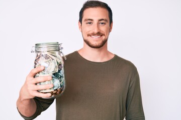 Young handsome man holding jar with savings looking positive and happy standing and smiling with a confident smile showing teeth