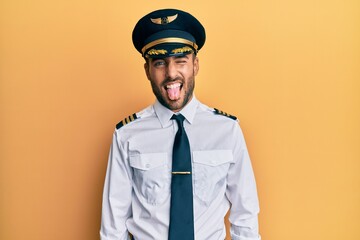 Handsome hispanic man wearing airplane pilot uniform sticking tongue out happy with funny expression. emotion concept.