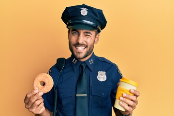 Handsome hispanic police man eating donut and drinking coffee winking looking at the camera with...