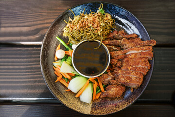Asian dish with roasted duck, vegetables, noodles and teriyaki sauce in a flat bowl on a wooden table, high angle view from above, selected focus