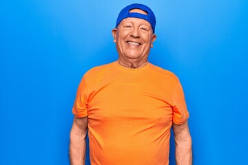 Fototapeta premium Senior handsome grey-haired man wearing casual t-shirt and cap over blue background with a happy and cool smile on face. Lucky person.