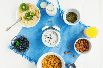 Time for breakfast.Top view alarm clock with healthy morning food. Bowl with cereals, nuts and berries, milk, fresh juice on blue napkin and white wooden table. Day diet planning and healthy eating.