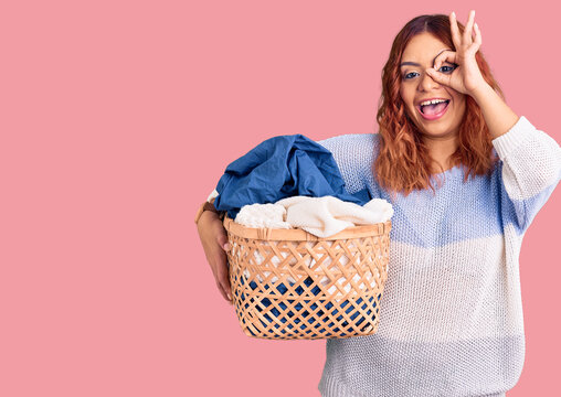 Young latin woman holding laundry basket smiling happy doing ok sign with hand on eye looking through fingers