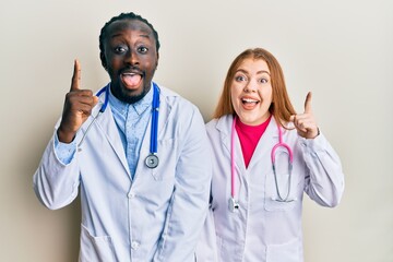 Young interracial couple wearing doctor uniform and stethoscope pointing finger up with successful...