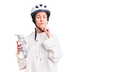 Beautiful brunette young woman wearing bike helmet and holding water bottle serious face thinking about question with hand on chin, thoughtful about confusing idea