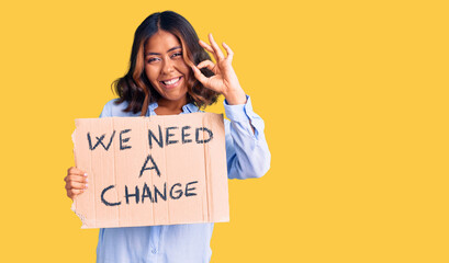 Young beautiful mixed race woman holding we need a change banner doing ok sign with fingers, smiling friendly gesturing excellent symbol