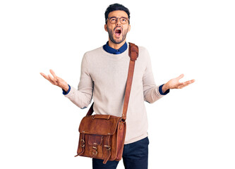 Young handsome hispanic man wearing leather bag crazy and mad shouting and yelling with aggressive expression and arms raised. frustration concept.