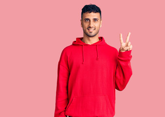 Young hispanic man wearing casual clothes smiling with happy face winking at the camera doing victory sign. number two.