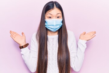 Young beautiful chinese woman wearing medical mask clueless and confused with open arms, no idea...