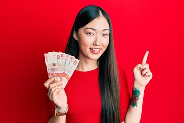 Young chinese woman holding 10 colombian pesos banknotes smiling happy pointing with hand and finger to the side