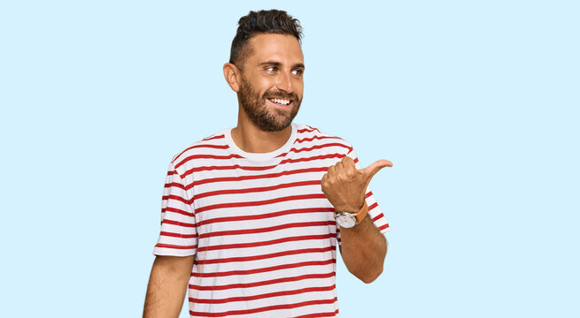 Handsome man with beard wearing striped tshirt smiling with happy face looking and pointing to the side with thumb up.
