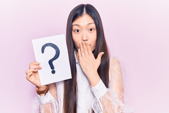 Young beautiful chinese woman holding question mark covering mouth with hand, shocked and afraid for mistake. surprised expression