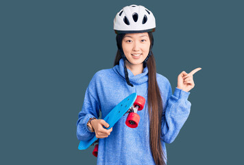 Young beautiful chinese woman wearing security helmet holding skate smiling happy pointing with hand and finger to the side