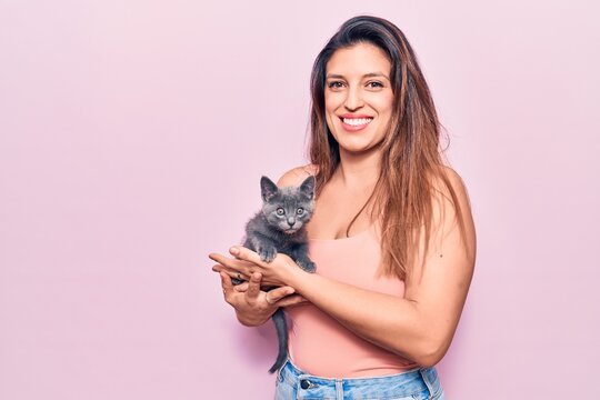 Young beautiful latin woman smilling happy. Standing with smile on face holding adorable cat over isolated pink background