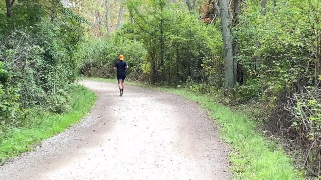 Rear view of man running on a dirt trail in the woods wearing an orange hat on his head backwards.