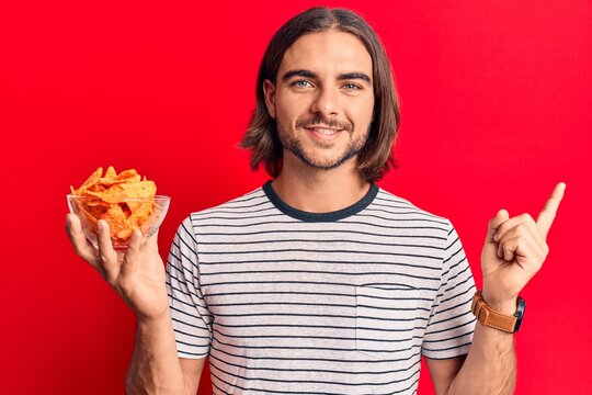 Young handsome man holding nachos potato chips smiling happy pointing with hand and finger to the side