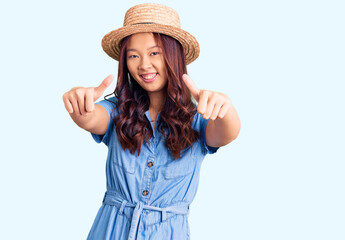 Young beautiful chinese girl wearing summer hat approving doing positive gesture with hand, thumbs up smiling and happy for success. winner gesture.
