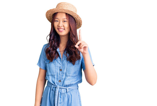 Young beautiful chinese girl wearing summer hat pointing finger up with successful idea. exited and happy. number one.