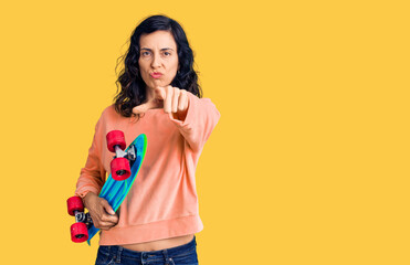 Young beautiful hispanic woman holding skate pointing with finger to the camera and to you, confident gesture looking serious