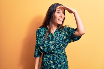 Young beautiful brunette woman wearing casual floral dress standing over yellow background very happy and smiling looking far away with hand over head. Searching concept.