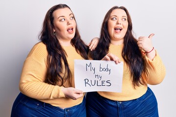 Young plus size twins holding my body my rules banner pointing thumb up to the side smiling happy with open mouth