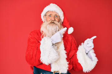 Old senior man with grey hair and long beard wearing traditional santa claus costume smiling and looking at the camera pointing with two hands and fingers to the side.