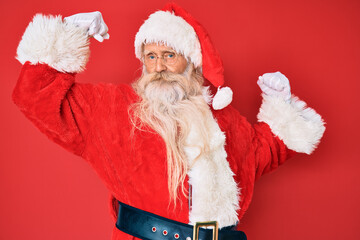 Old senior man with grey hair and long beard wearing traditional santa claus costume showing arms muscles smiling proud. fitness concept.