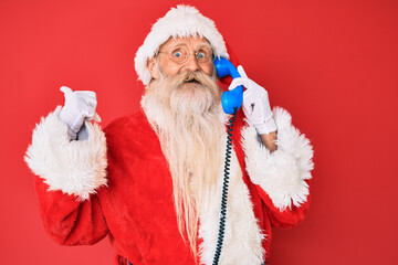 Old senior man wearing santa claus costume using vintage telephone pointing thumb up to the side smiling happy with open mouth