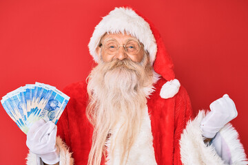 Old senior man with grey hair and long beard wearing santa claus costume holding african rands screaming proud, celebrating victory and success very excited with raised arm