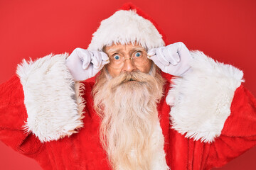 Old senior man with grey hair and long beard wearing santa claus costume holding glasses in shock face, looking skeptical and sarcastic, surprised with open mouth