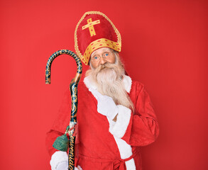 Old senior man with grey hair and long beard wearing traditional saint nicholas costume cheerful with a smile on face pointing with hand and finger up to the side with happy and natural expression