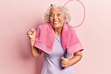 Senior grey-haired woman holding badminton racket wearing towel screaming proud, celebrating victory and success very excited with raised arm - obrazy, fototapety, plakaty