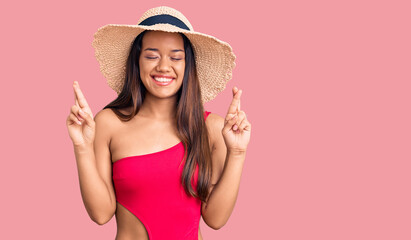 Young beautiful latin girl wearing swimwear and summer hat gesturing finger crossed smiling with hope and eyes closed. luck and superstitious concept.