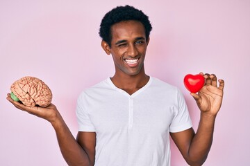 Handsome african man holding brain and heart winking looking at the camera with sexy expression,...