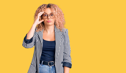 Young blonde woman with curly hair wearing business jacket and glasses worried and stressed about a problem with hand on forehead, nervous and anxious for crisis