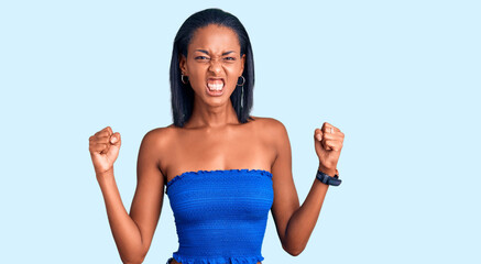 Young african american woman wearing casual summer clothes angry and mad raising fists frustrated...
