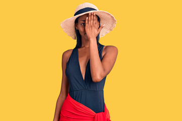 Young african american woman wearing swimsuit and summer hat covering one eye with hand, confident smile on face and surprise emotion.