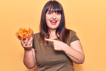 Young plus size woman holding nachos potato chips smiling happy pointing with hand and finger